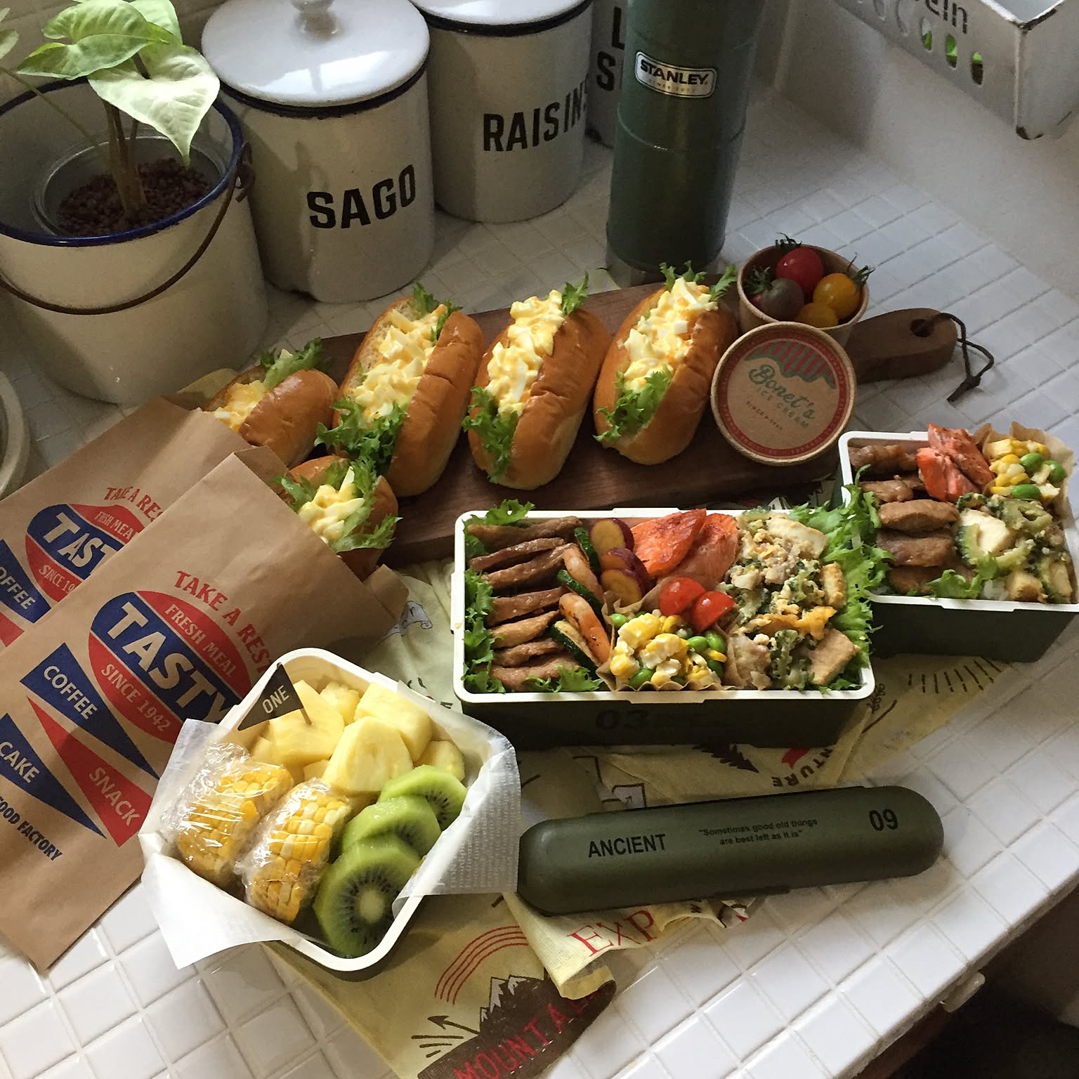 Two packed rectangular adult bento boxes with sandwiches, fruit, cutlery, and a drink bottle on a kitchen counter.