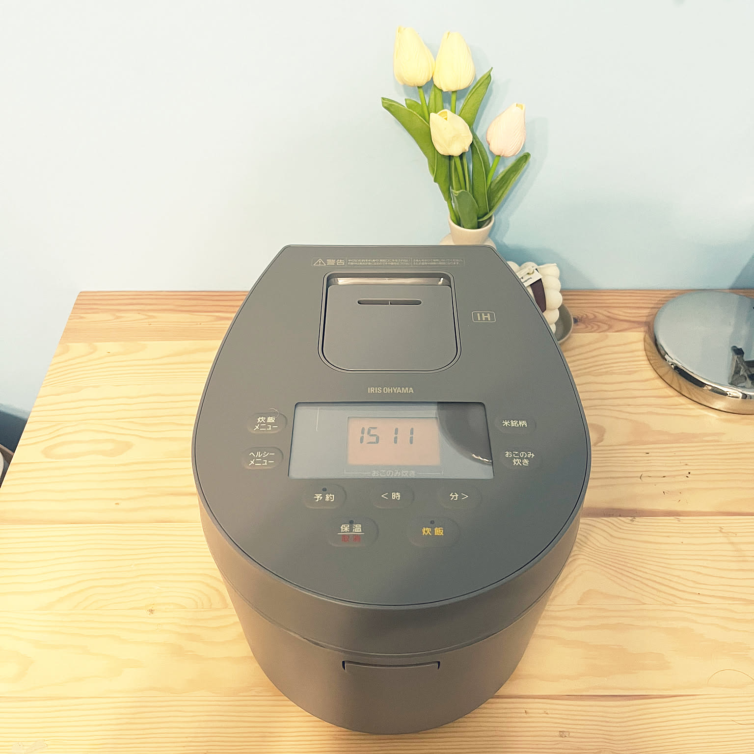 A gray Iris Ohyama three-cup IH rice cooker on a wooden table with flowers behind it.