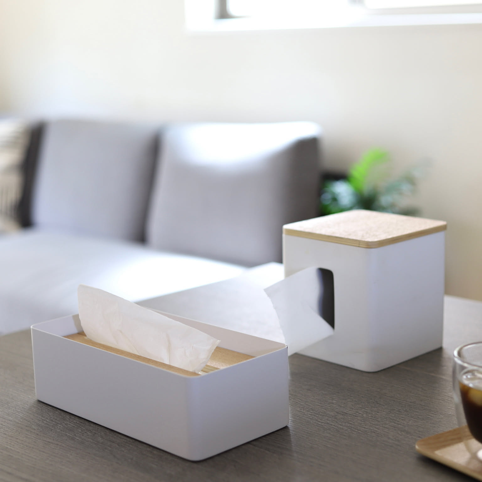 Two white steel tissue cases with wood lids on a living-room table, with a gray sofa in the background