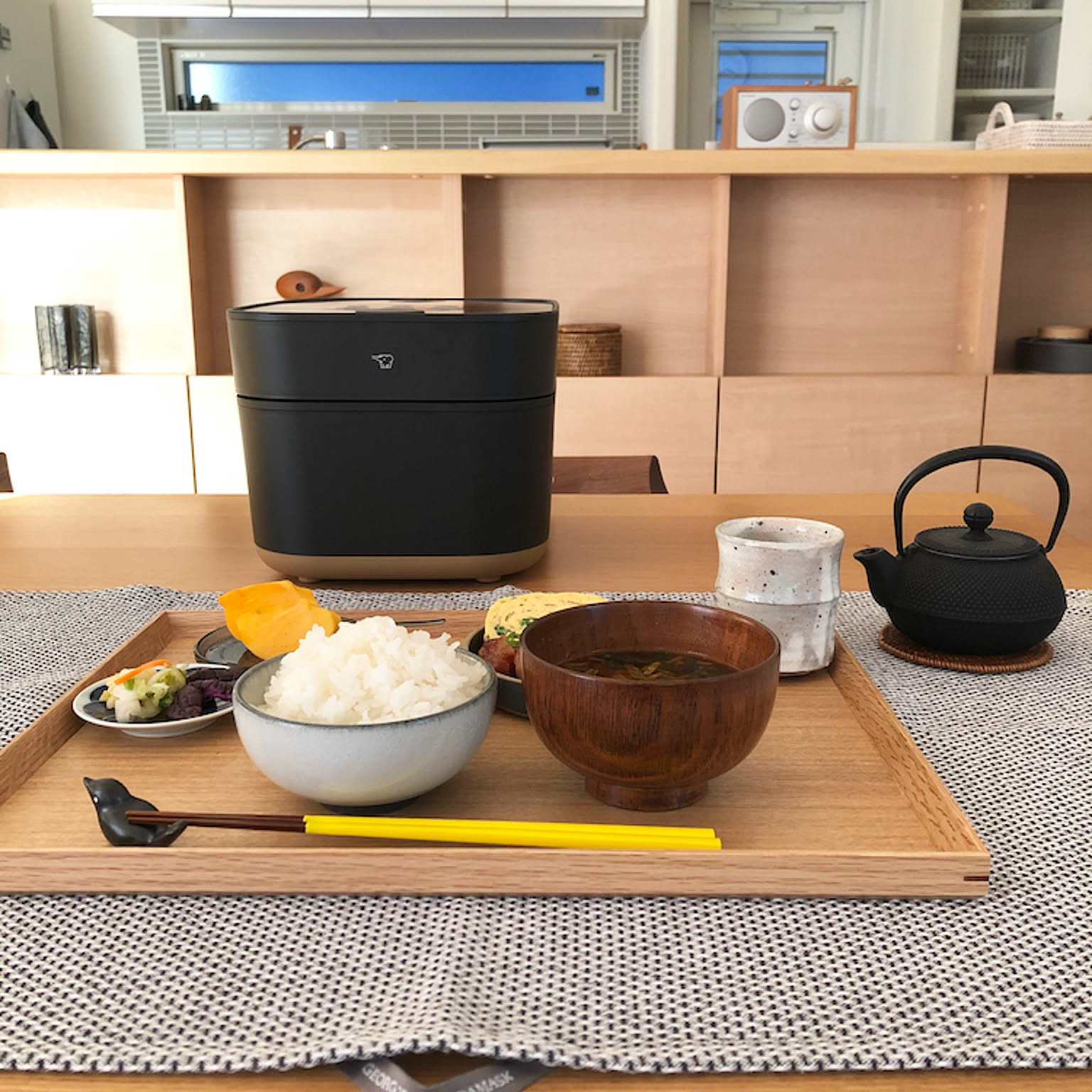A Zojirushi STAN. rice cooker behind a wooden tray holding a bowl of freshly cooked rice, miso soup, side dishes, chopsticks, and a cast-iron teapot.