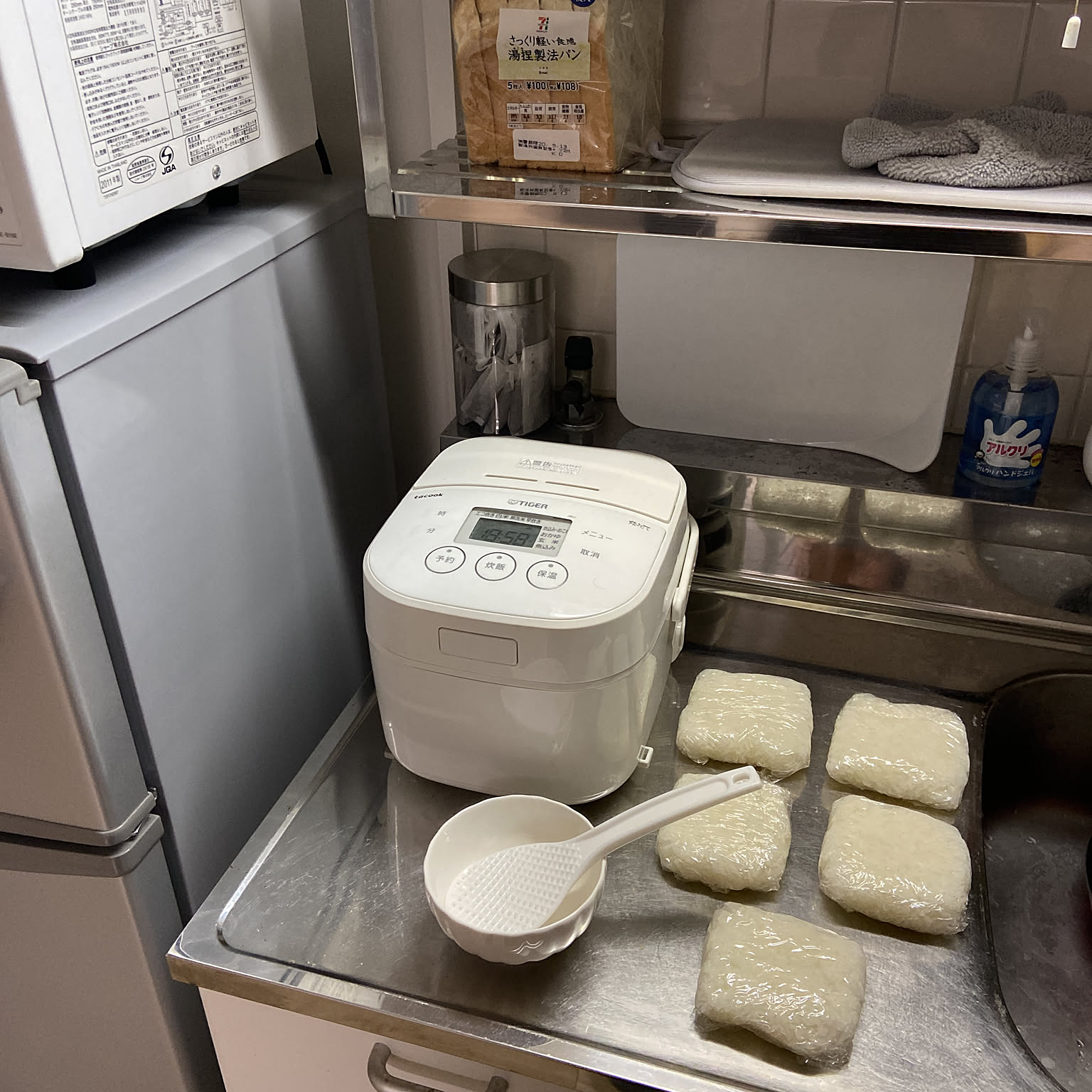 A compact rice cooker beside five single-portion rice packs wrapped on a stainless counter.