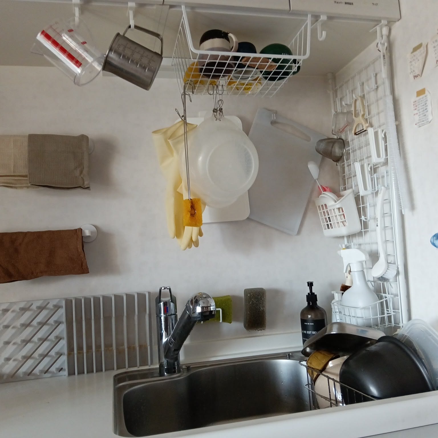A compact Japanese sink area with a water-draining basket, hanging cups, gloves, cutting boards, and small tools stored around the sink wall.