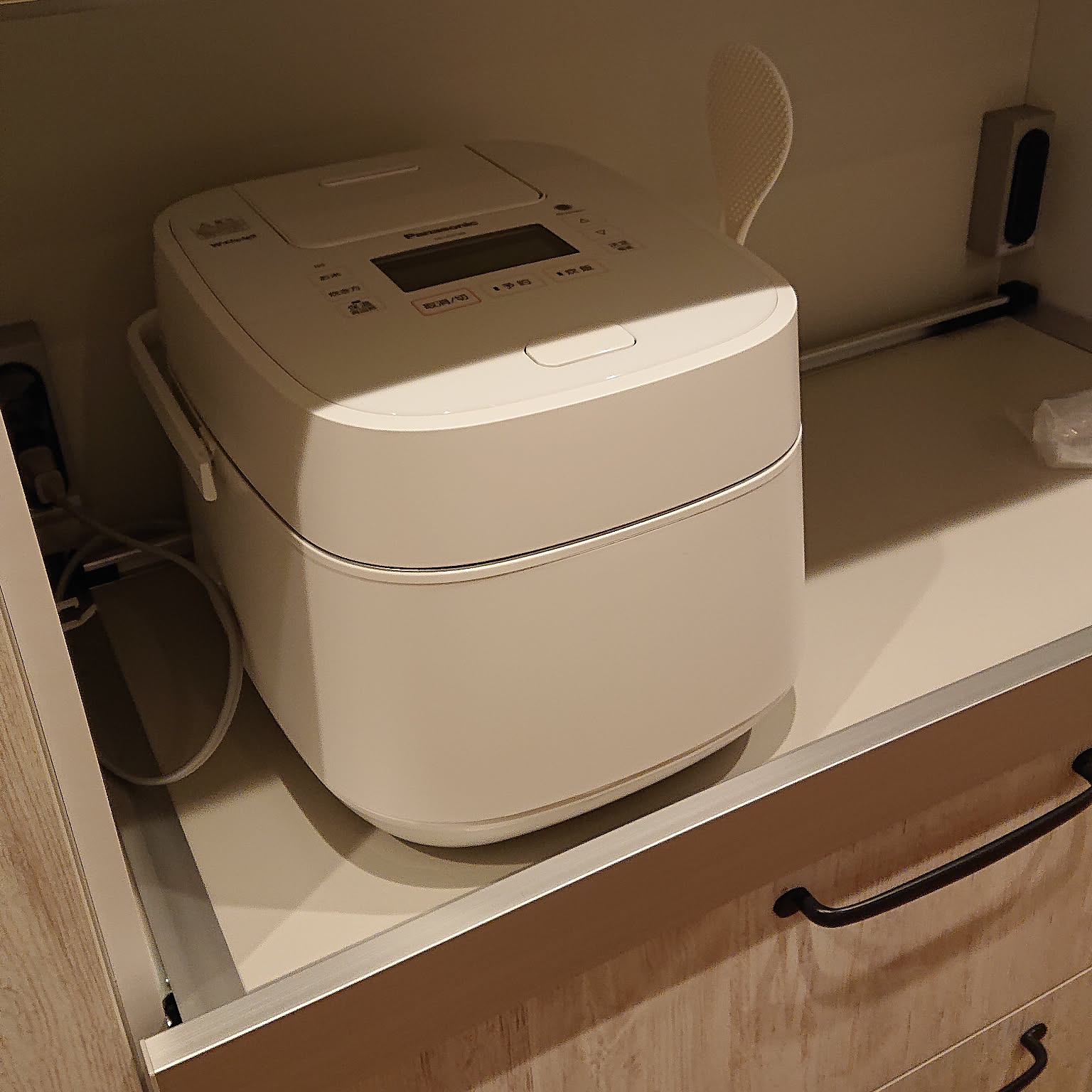 A white Panasonic rice cooker on a pulled-out kitchen shelf with its power cord running down behind the drawer