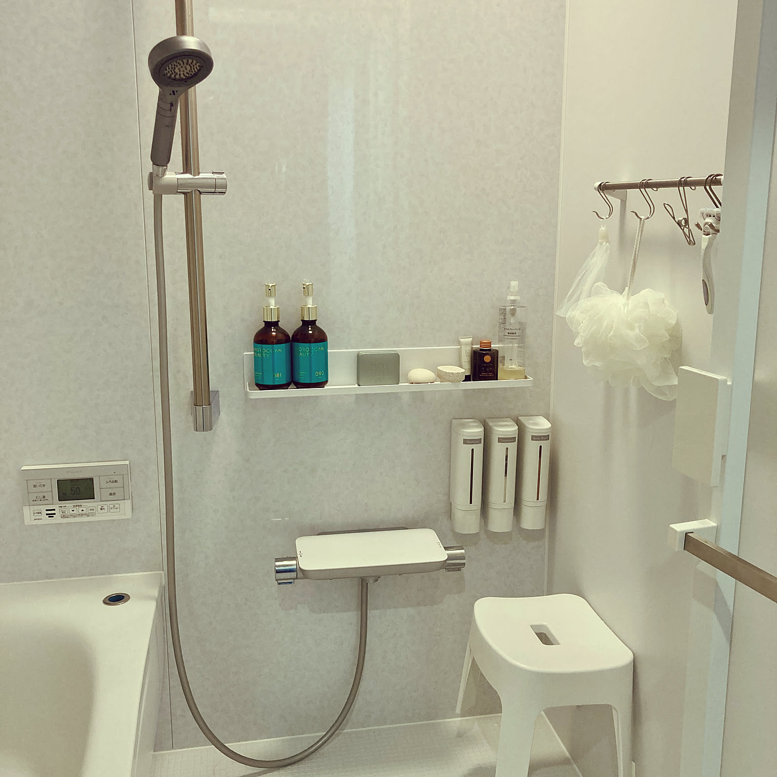 A compact white Japanese bathroom with wall-mounted dispensers, hanging bath puffs, a narrow shelf, and a clear floor.