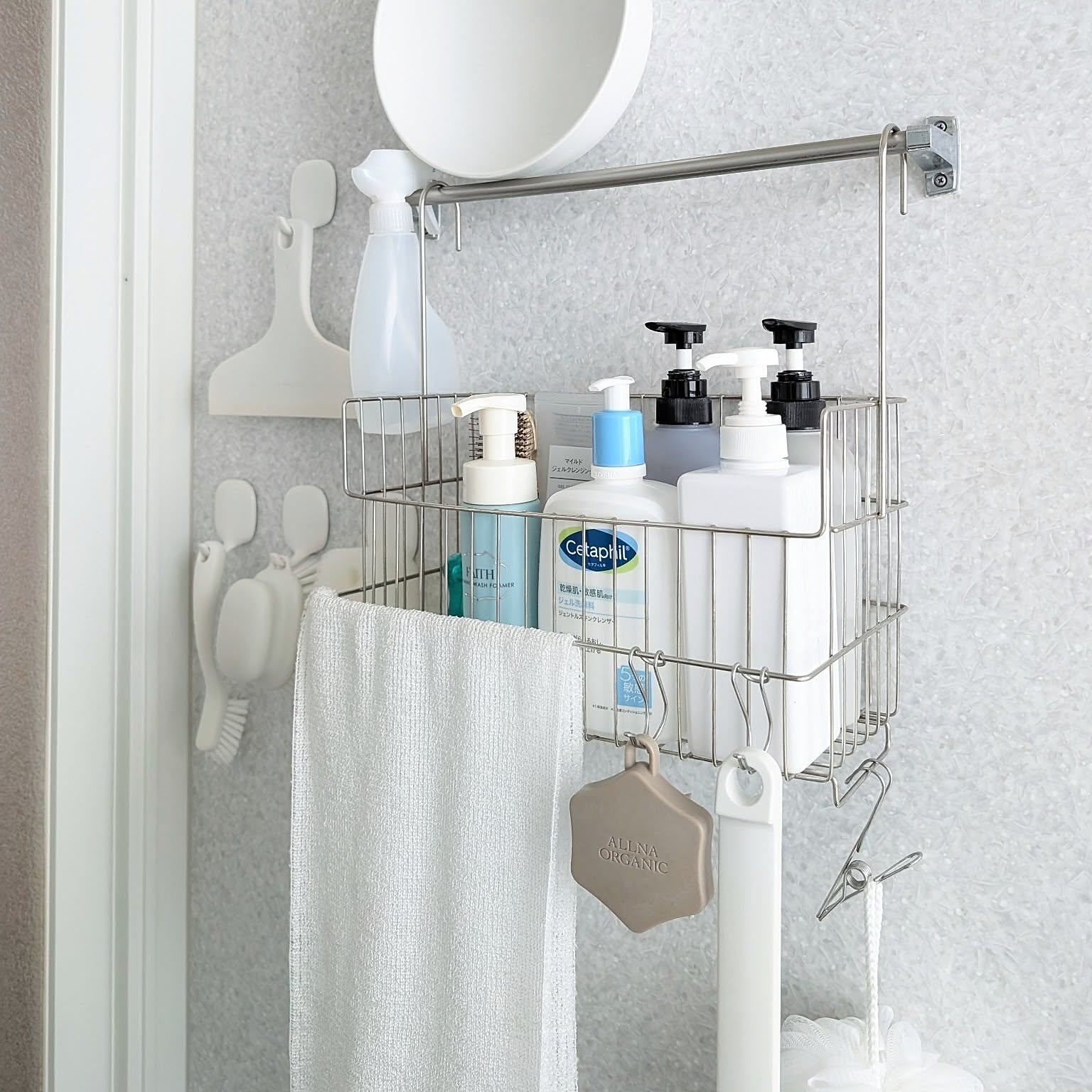 A stainless wire bathroom basket hanging from a rail with bottles, soap, a towel, and small tools inside.