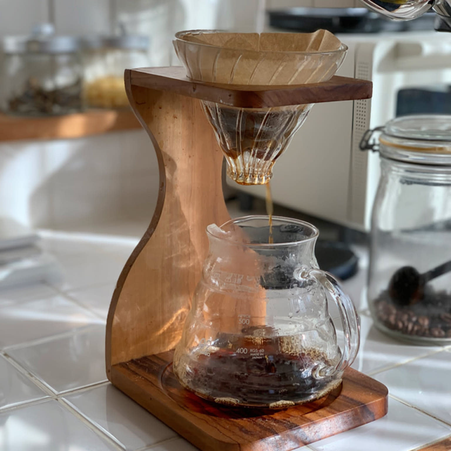 A HARIO V60 dripper in an olive-wood stand brewing into a glass server in a bright kitchen.