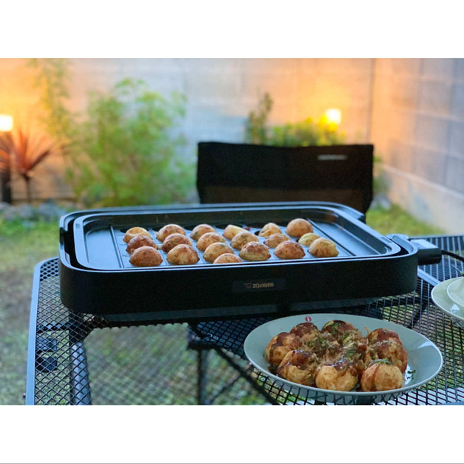 A black electric hot plate with takoyaki on a table and a plate of cooked takoyaki nearby.