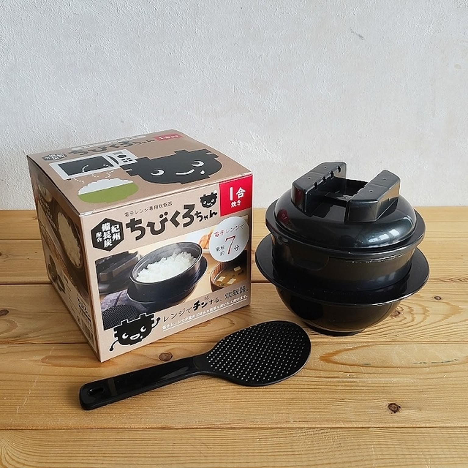 A one-go microwave rice cooker with its package and black rice paddle on a wooden table.