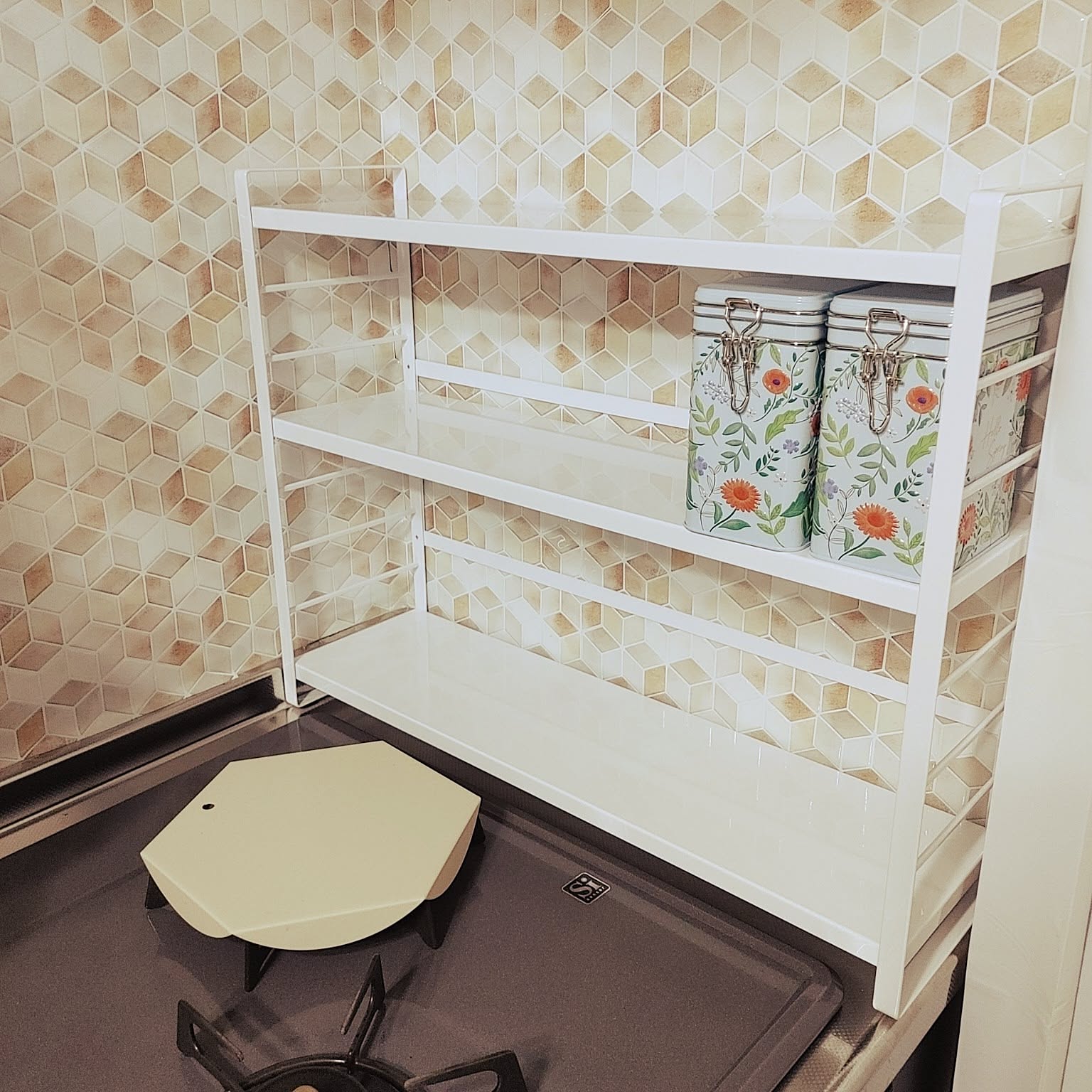 A white Yamazaki Tower countertop three-shelf rack placed beside a stove and holding small storage tins.