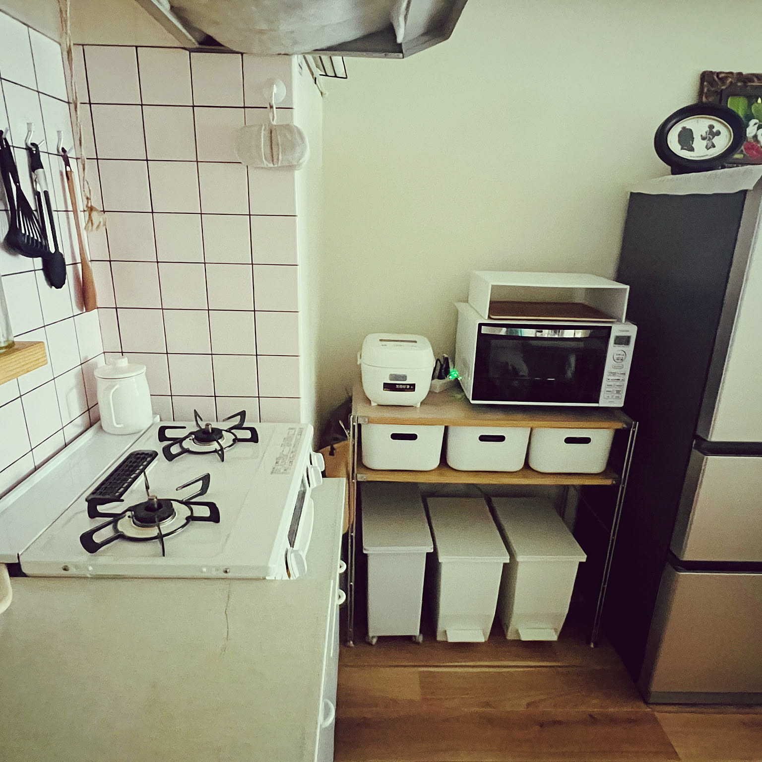 A narrow Japanese rental kitchen with a gas stove, microwave, rice cooker, refrigerator, and simple white storage boxes used to keep the small cooking area workable.