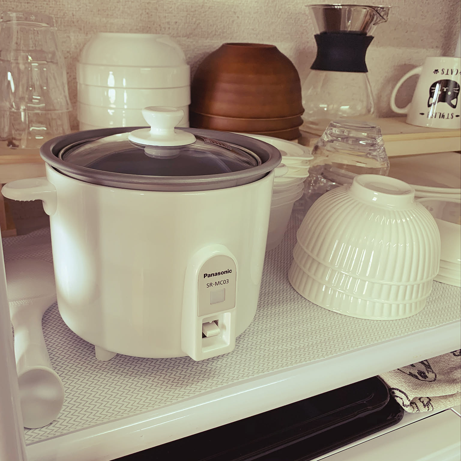 A white Panasonic mini rice cooker stored on a shelf beside bowls and coffee gear.