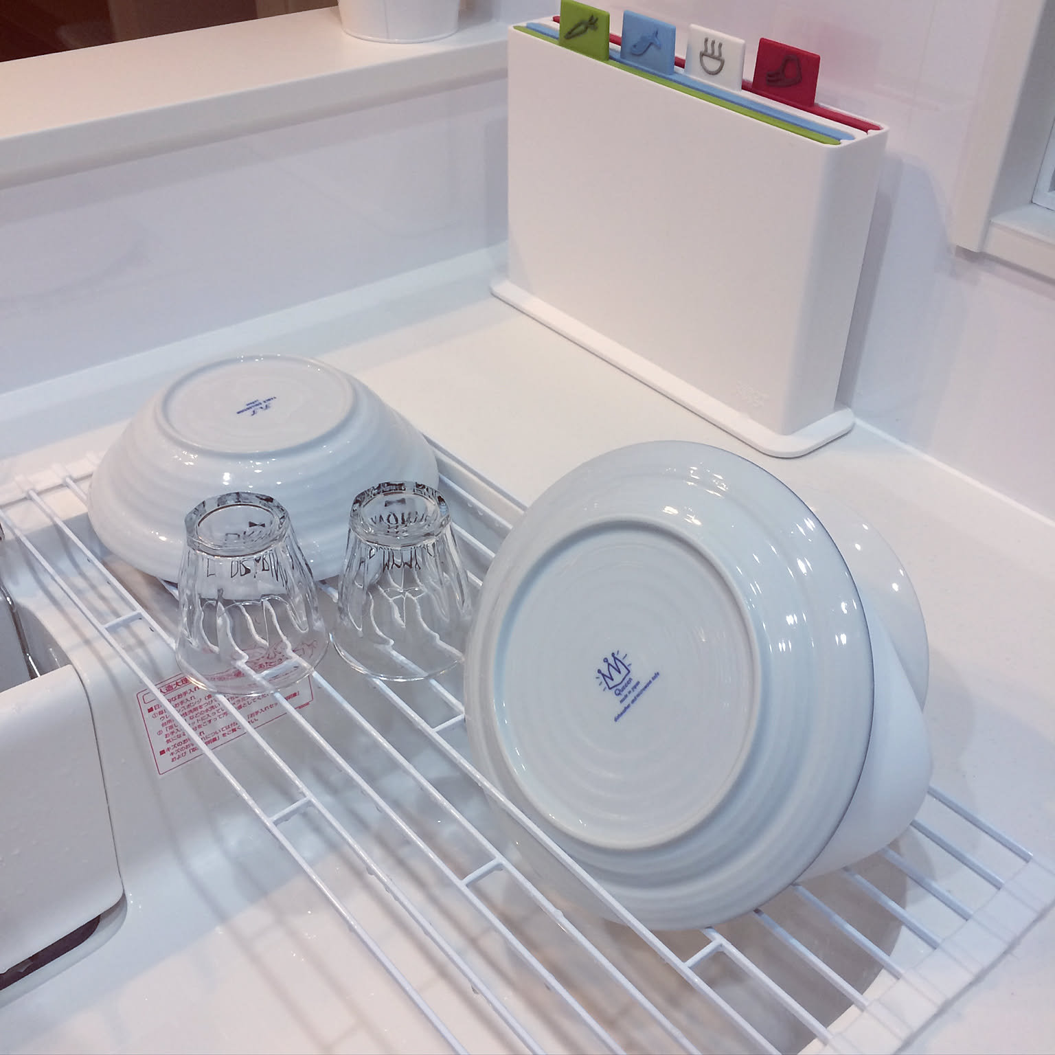 A white Yamazaki Tower folding dish drainer placed across a kitchen sink with plates and glasses drying on it.