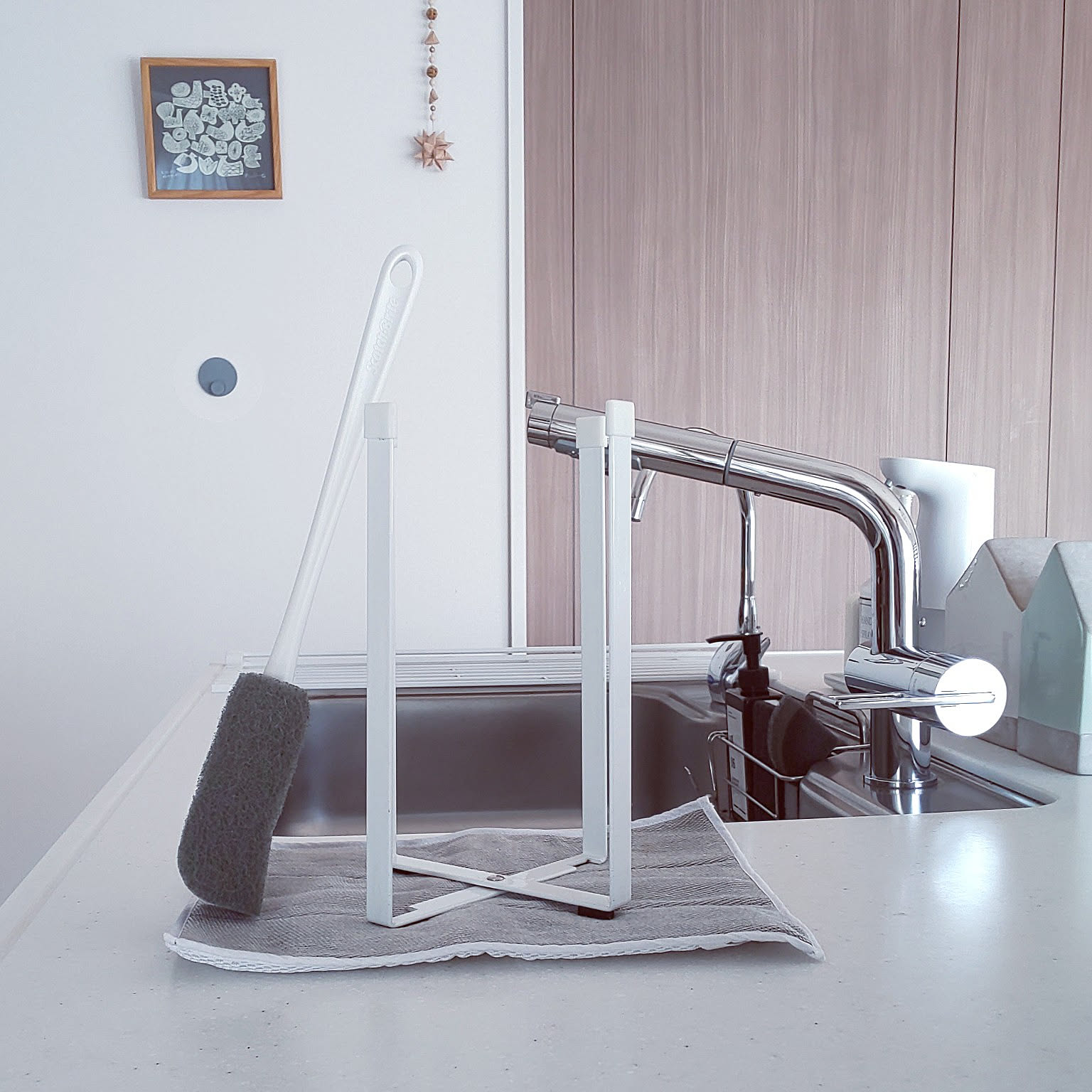 A folding white steel bag stand beside a kitchen sink, drying a long bottle brush and a dishcloth