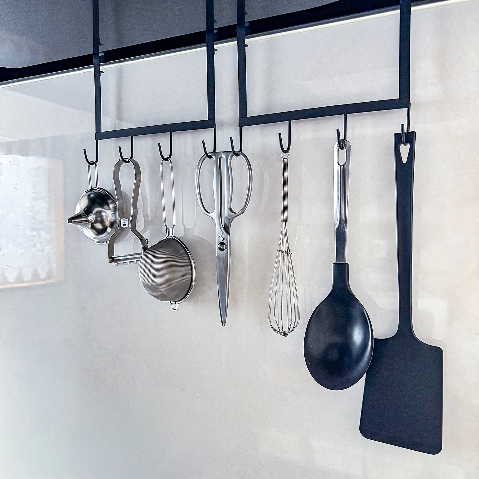 A close-up of ladles, scissors, peelers, strainers, and spatulas hanging from black hooks under a kitchen vent hood.