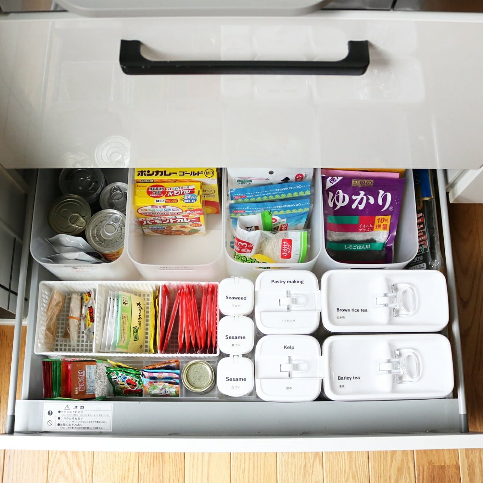 A kitchen stock drawer with open white bins separating cans, tea, retort food, soup packets, and snacks.