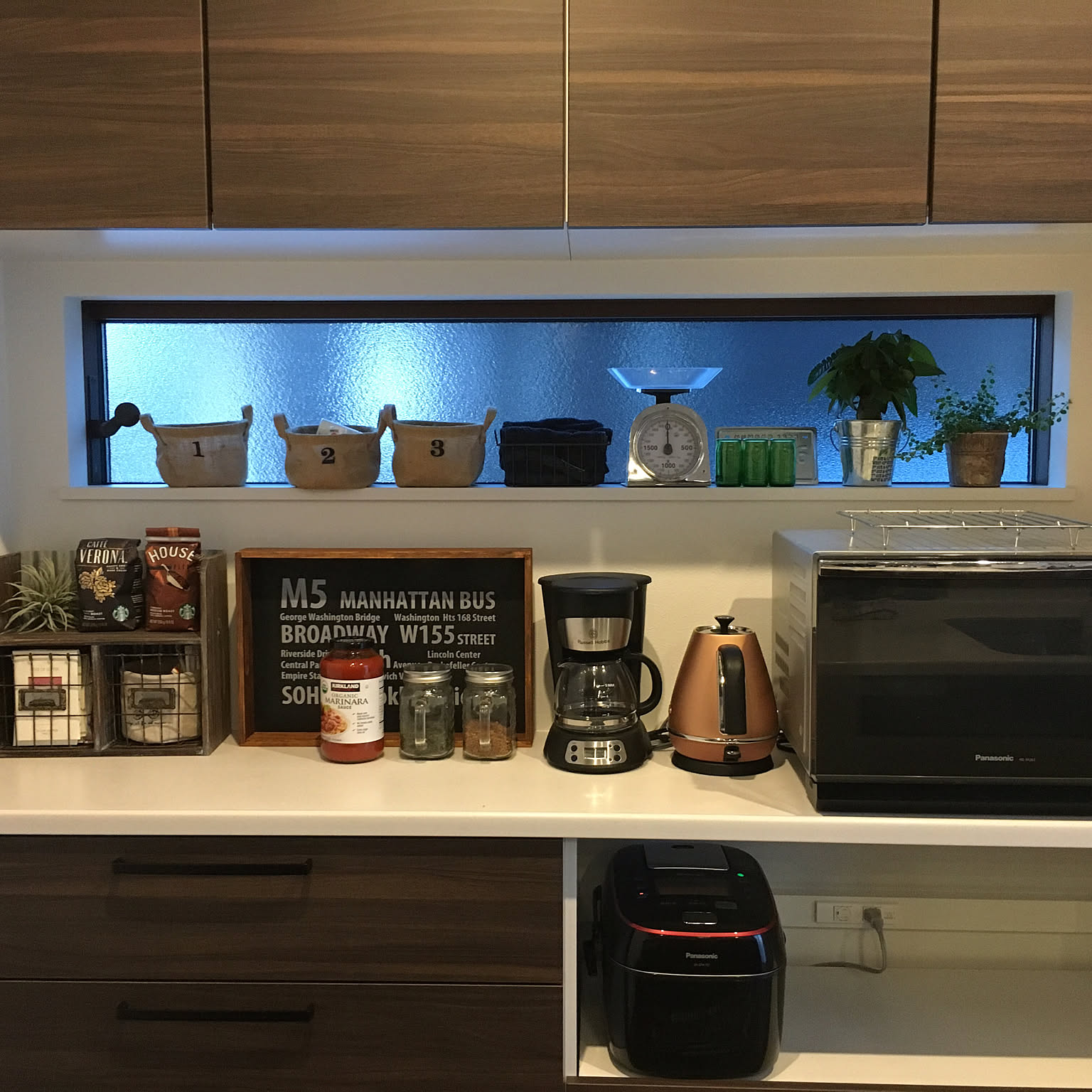 A Japanese kitchen appliance counter with a black Panasonic pressure-IH rice cooker on a pull-out shelf below a toaster oven, coffee maker, and copper kettle