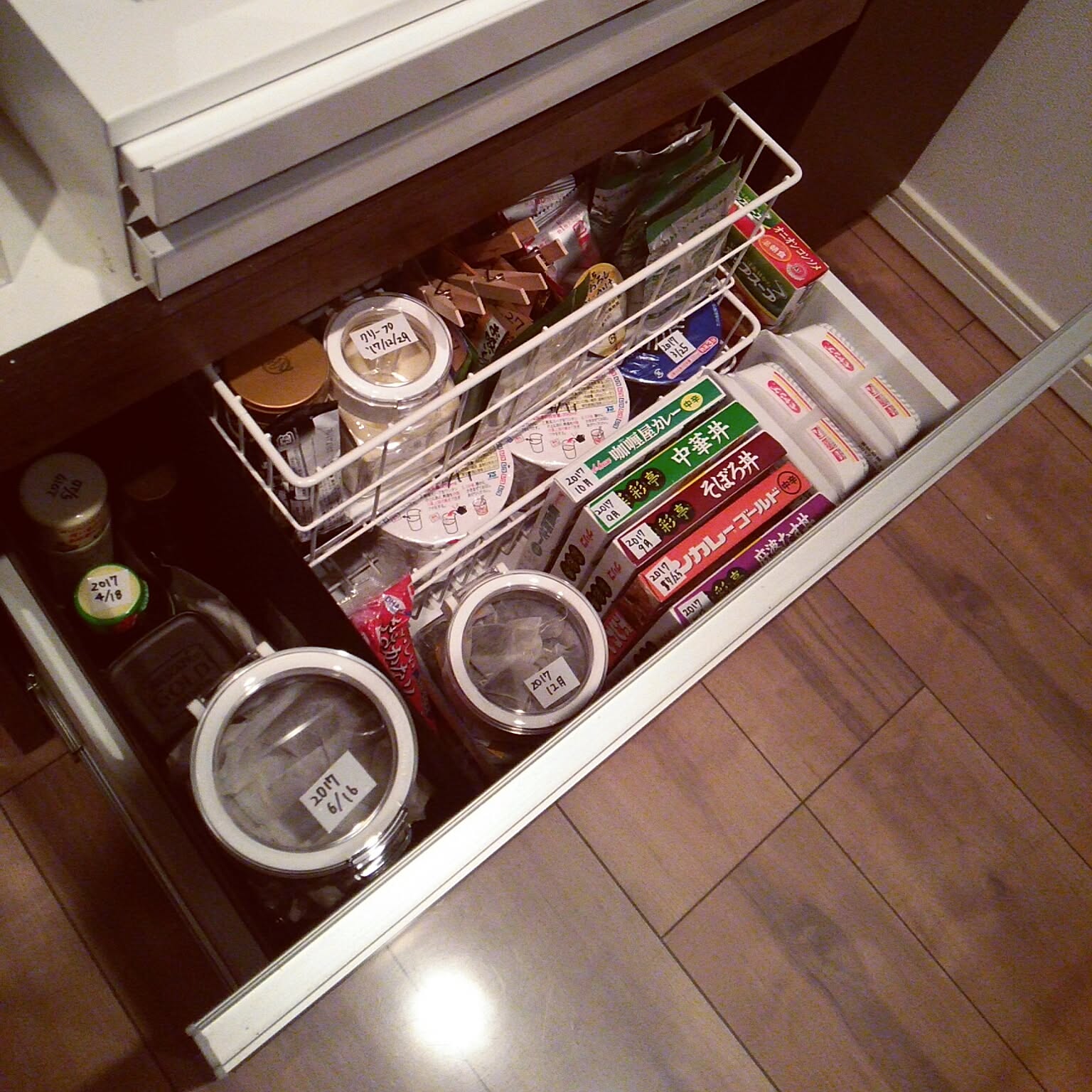 A pull-out kitchen cabinet drawer with wire baskets storing tea, packets, jars, and dry goods.