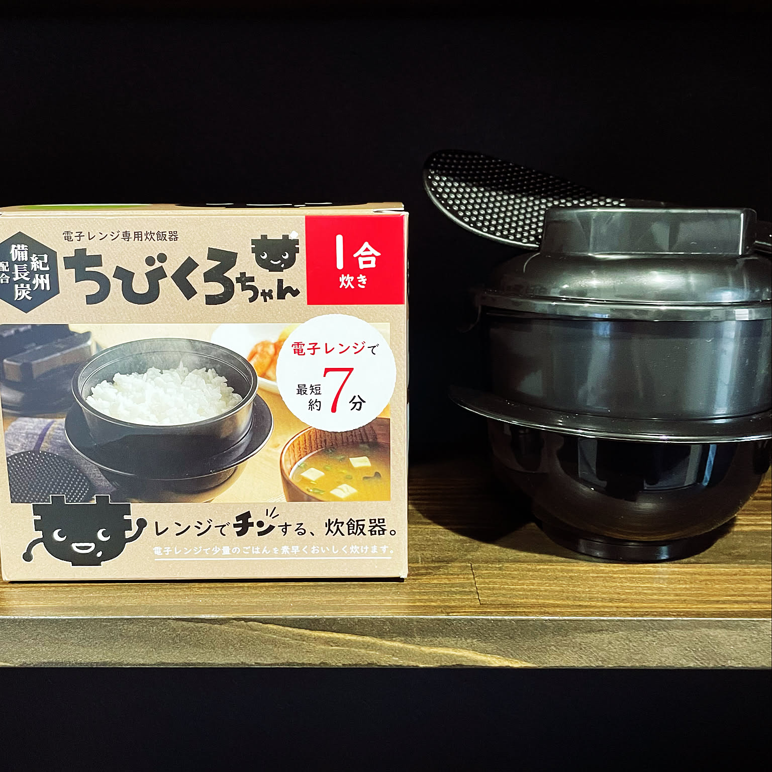 A black one-go microwave rice cooker next to its box and rice paddle.