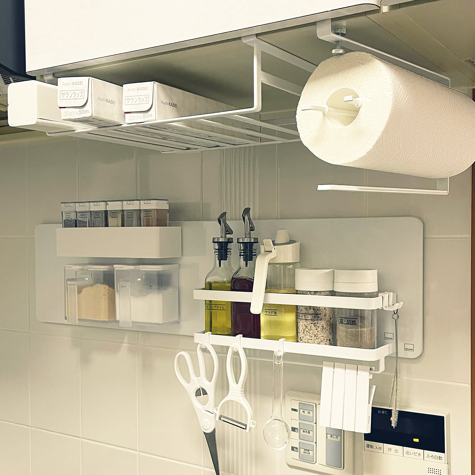 White steel under-shelf racks holding plastic wrap and a paper towel roll, above magnetic wall panels with seasoning bottles and tools