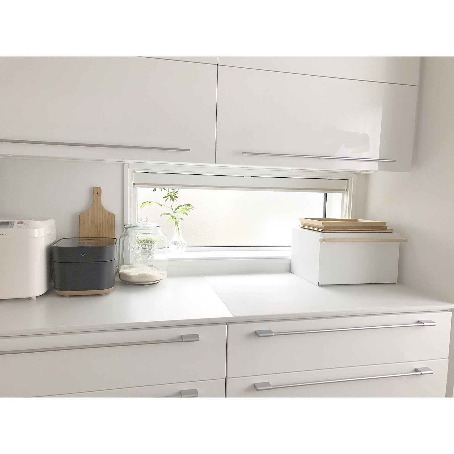 A white steel bread case with a wood lid on a tidy white kitchen counter next to a bread maker and a glass rice jar