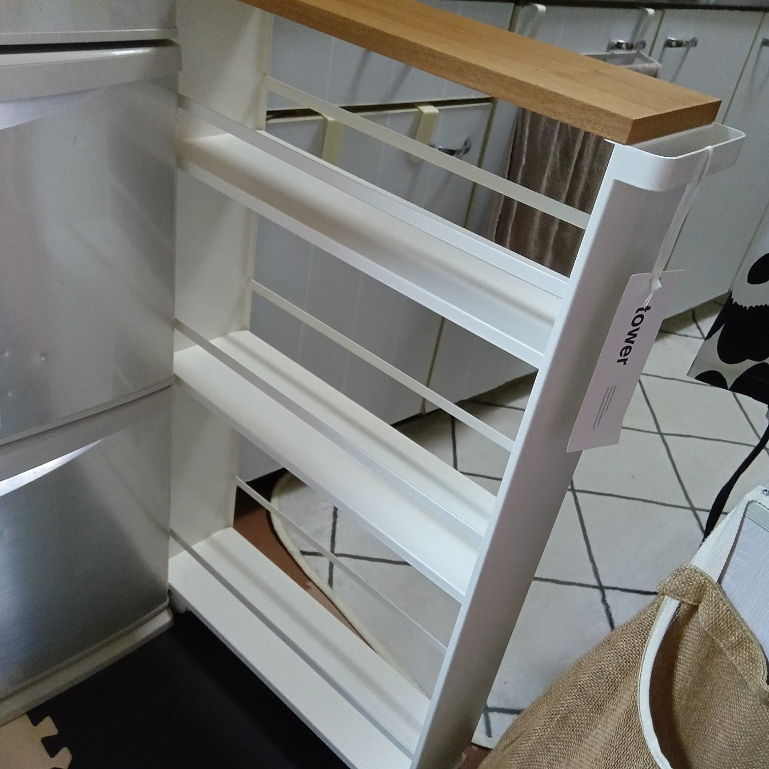 A white Yamazaki Tower rolling slim storage cart with a wood handle tucked into a narrow gap beside a refrigerator.