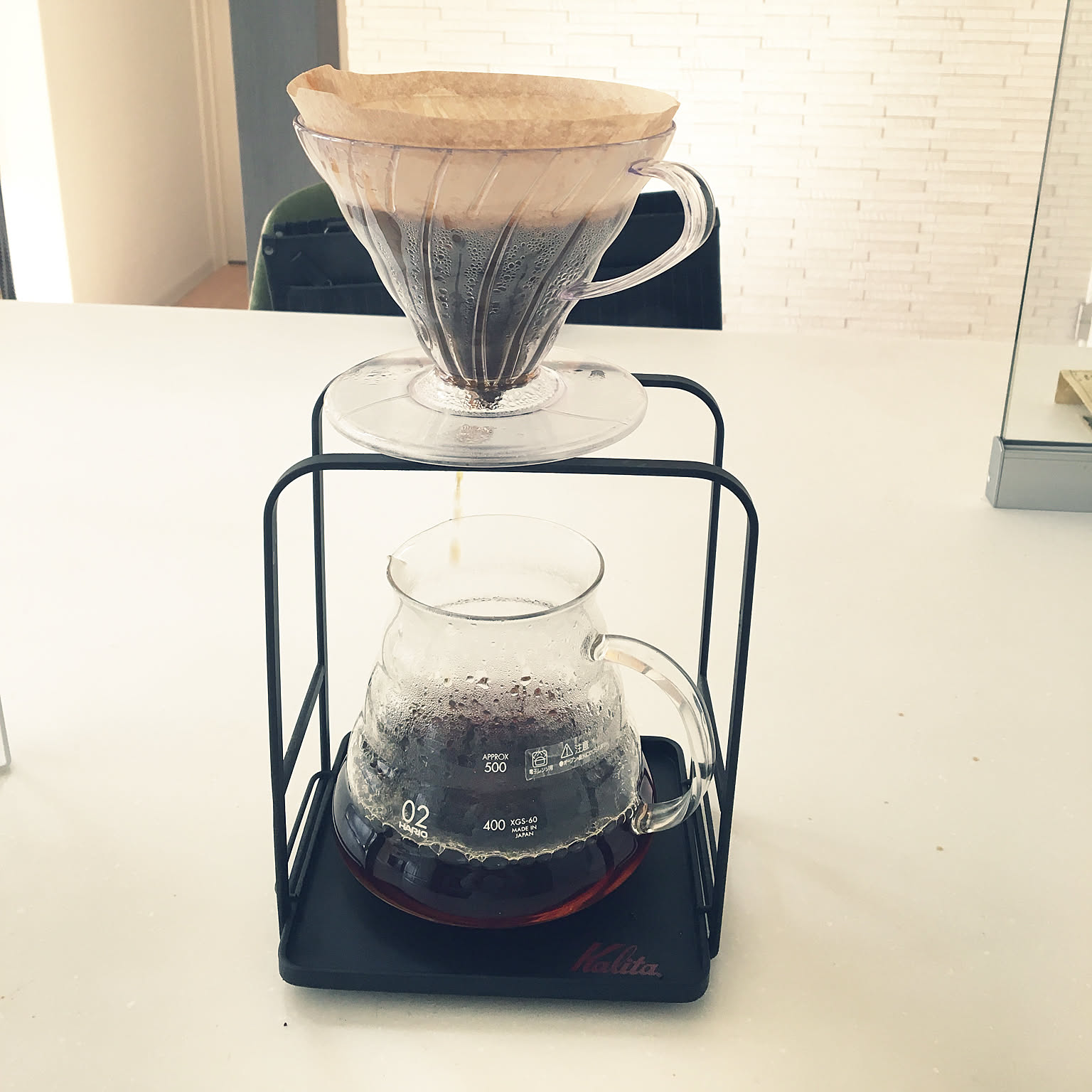 A clear V60 dripper brewing coffee into a glass server on a stand.