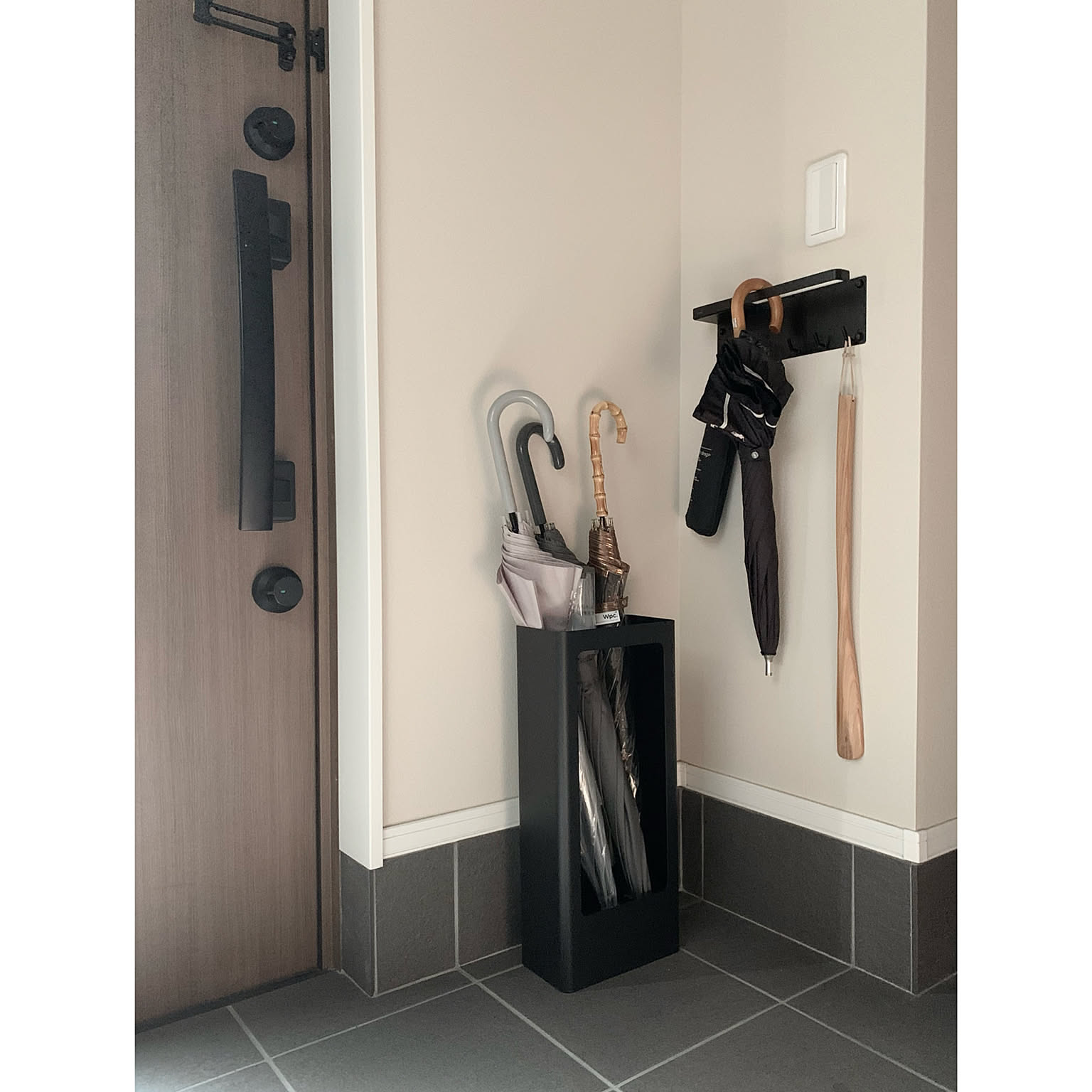 A small Japanese entryway with a slim black steel umbrella stand in the corner and a black wall hook holding a folded umbrella and shoehorn