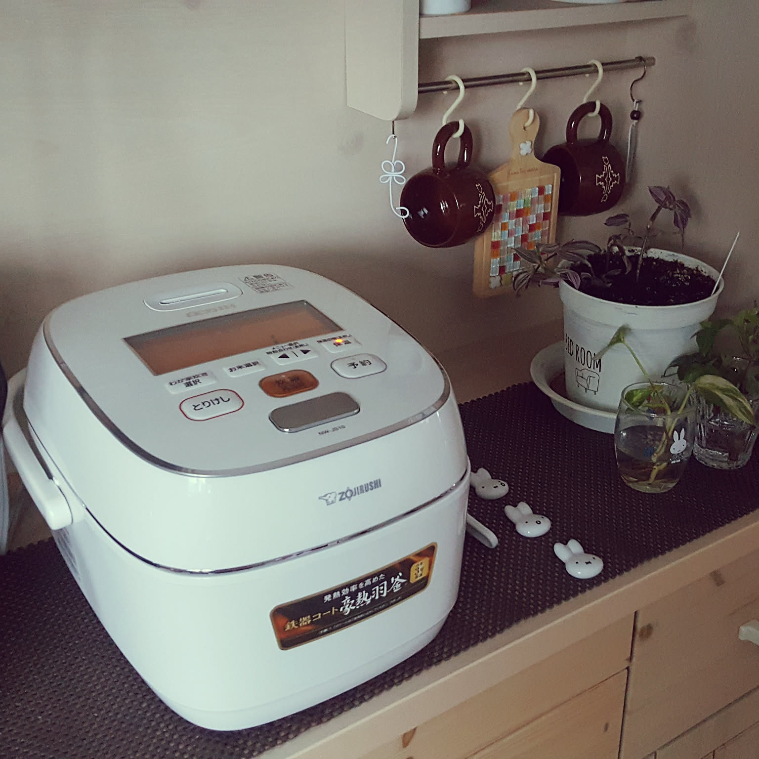 A white Zojirushi pressure-IH rice cooker with the ZOJIRUSHI wordmark on the front, on a kitchen shelf with mugs and a cutting board hanging above