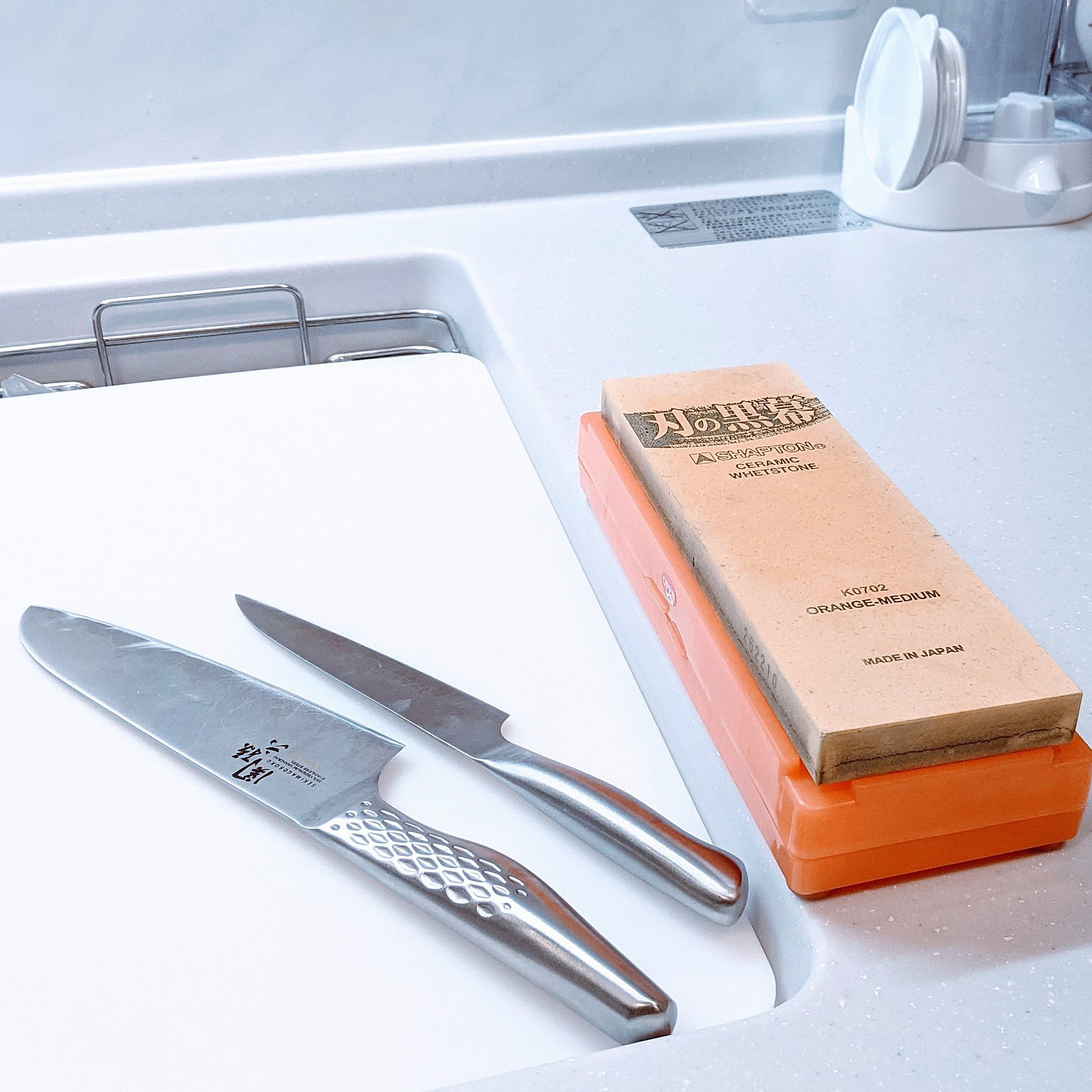 Two Japanese kitchen knives on a white board beside a whetstone near the sink.