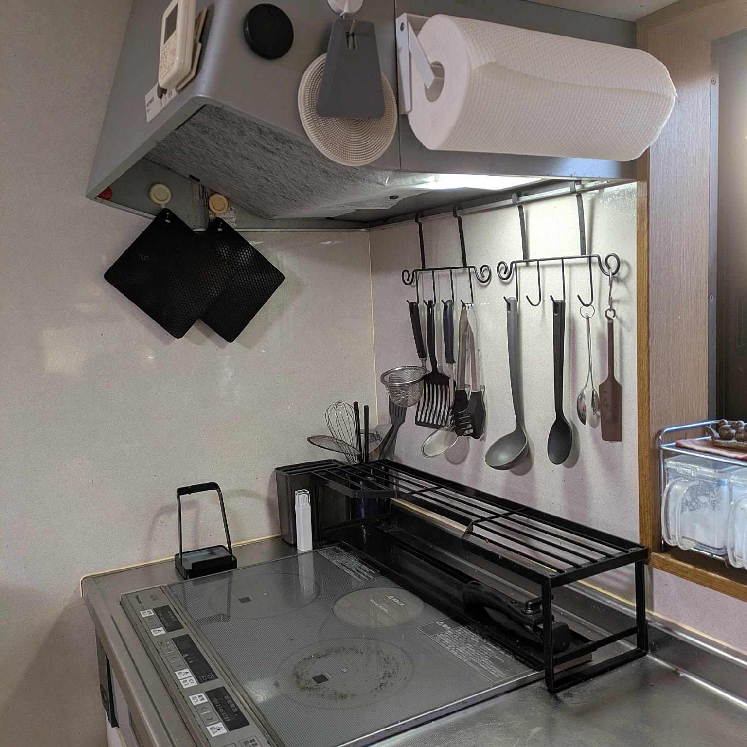 A black Yamazaki Tower cooktop rack and white paper towel holder arranged around an IH cooktop with hanging utensils.