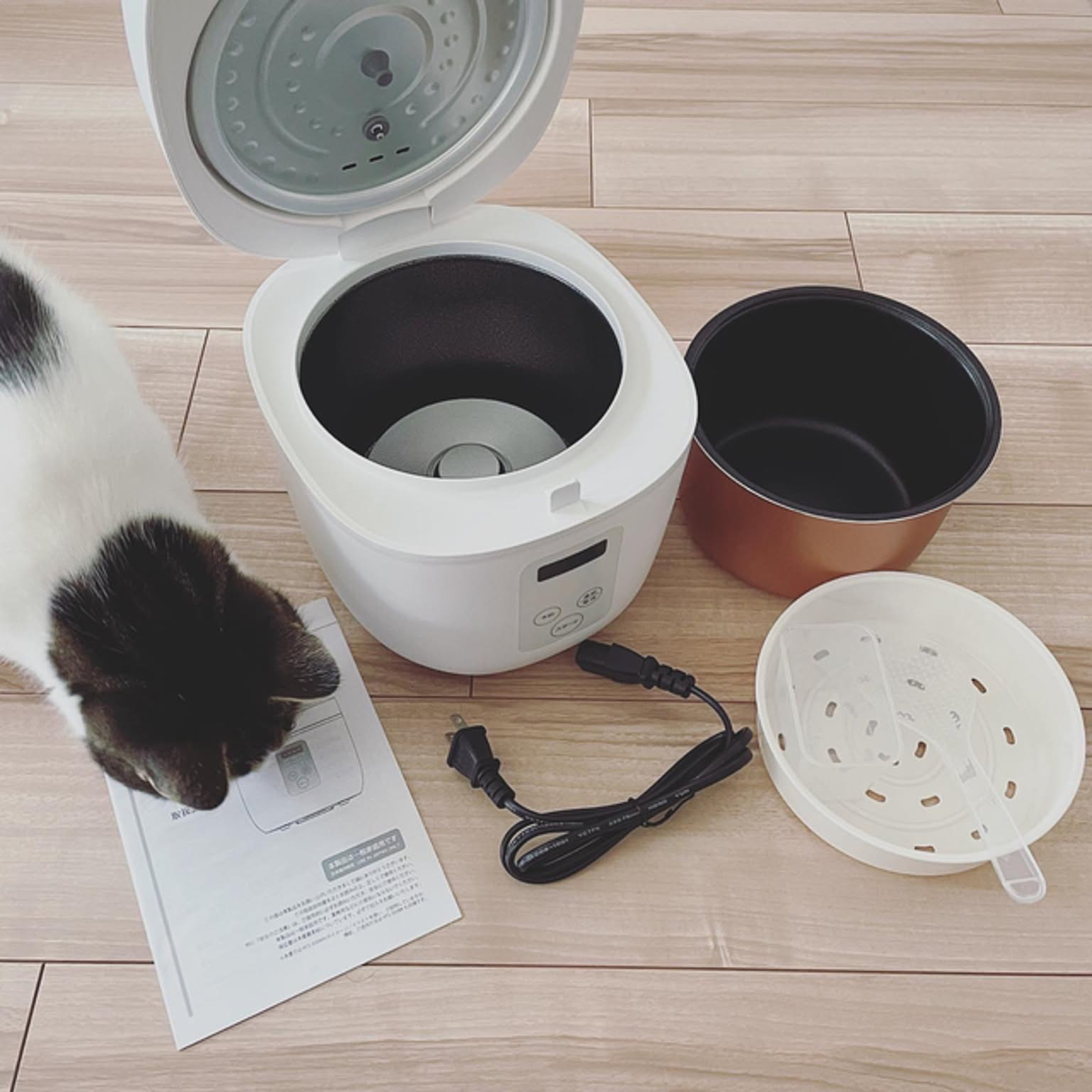 A white countertop rice cooker shown with its measuring cup, rice paddle, and power cord in a Japanese home.