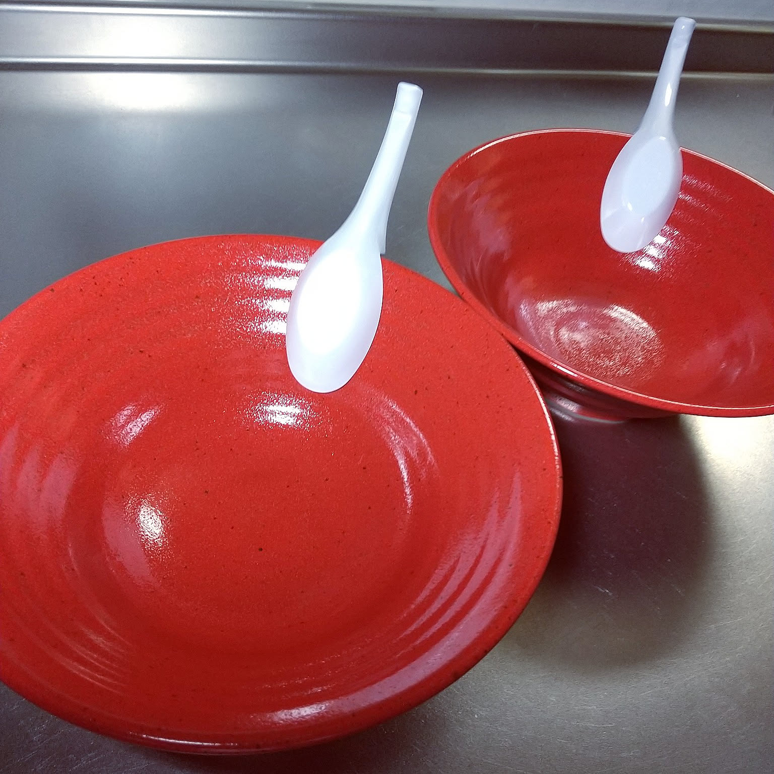 Two wide red ramen bowls with white soup spoons.