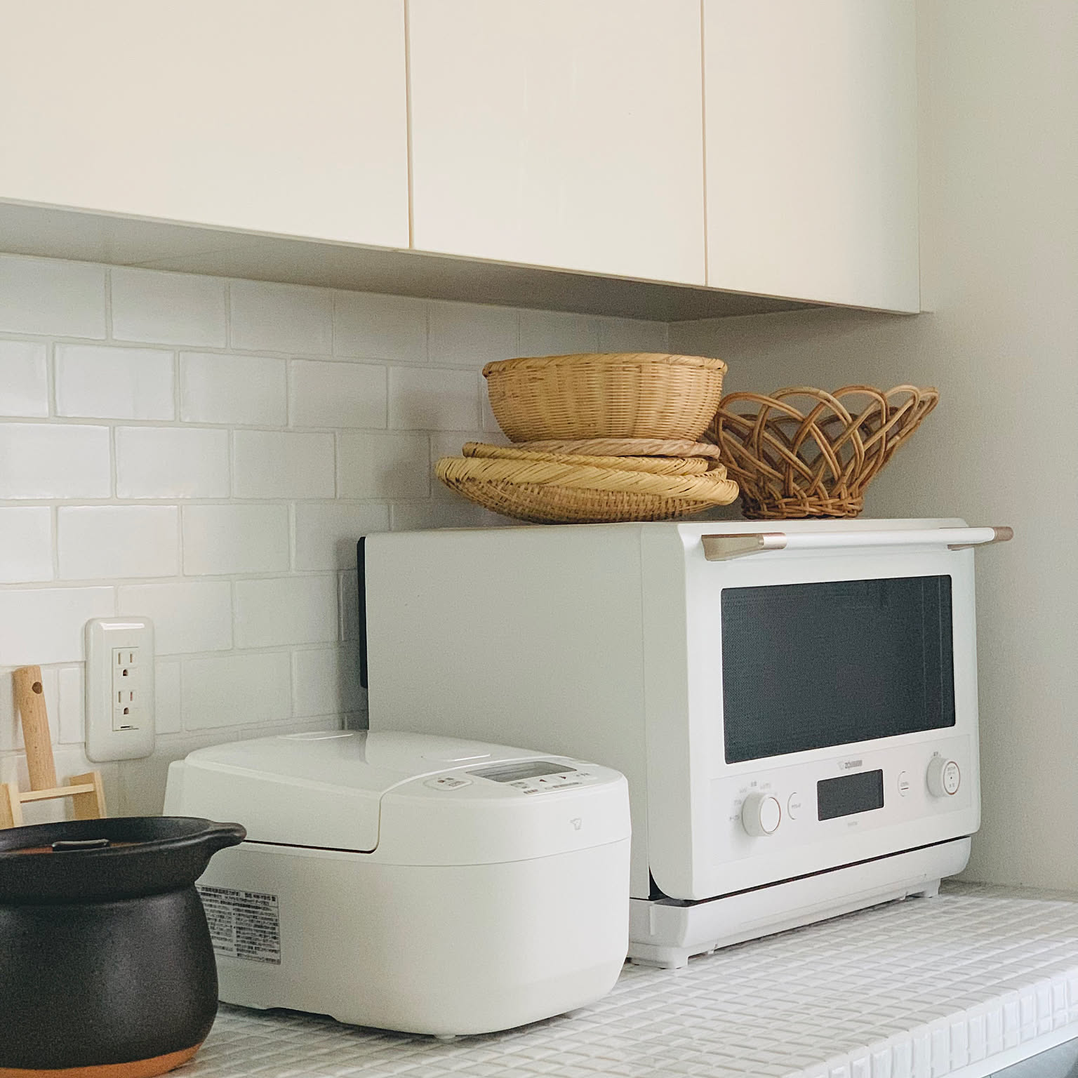 A white Zojirushi rice cooker next to a matching oven and a traditional Japanese donabe clay pot on a tiled kitchen counter.