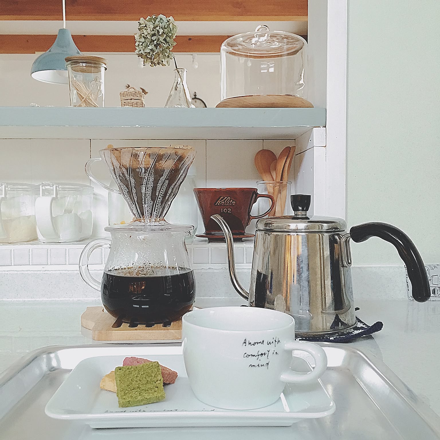 A clear HARIO V60 dripper brewing coffee into a glass server beside a stainless gooseneck kettle.