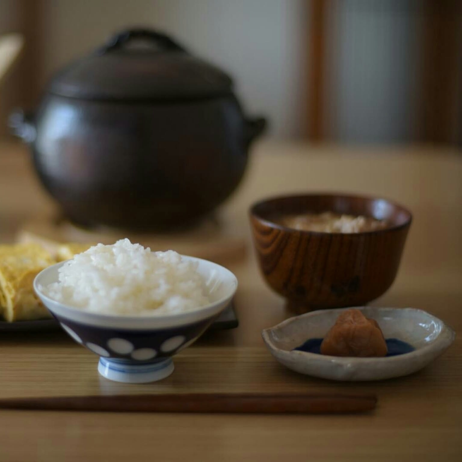Japanese table setting with rice bowl, soup, small dish, chopsticks, and a clay pot in the background.