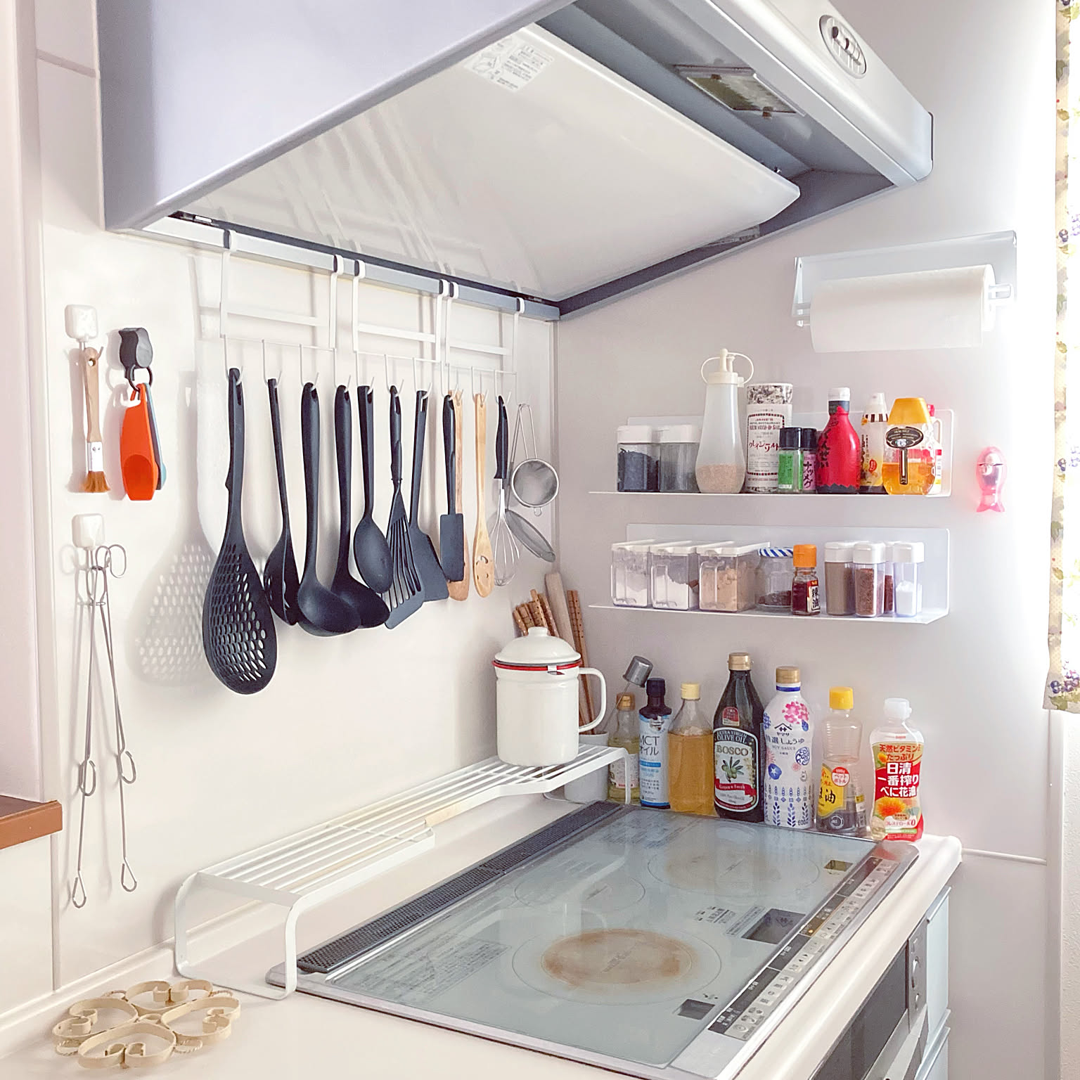 A compact Japanese kitchen with utensils on hood hooks, magnetic spice shelves on the wall, and a shelf-topped cover over the stove's rear vent