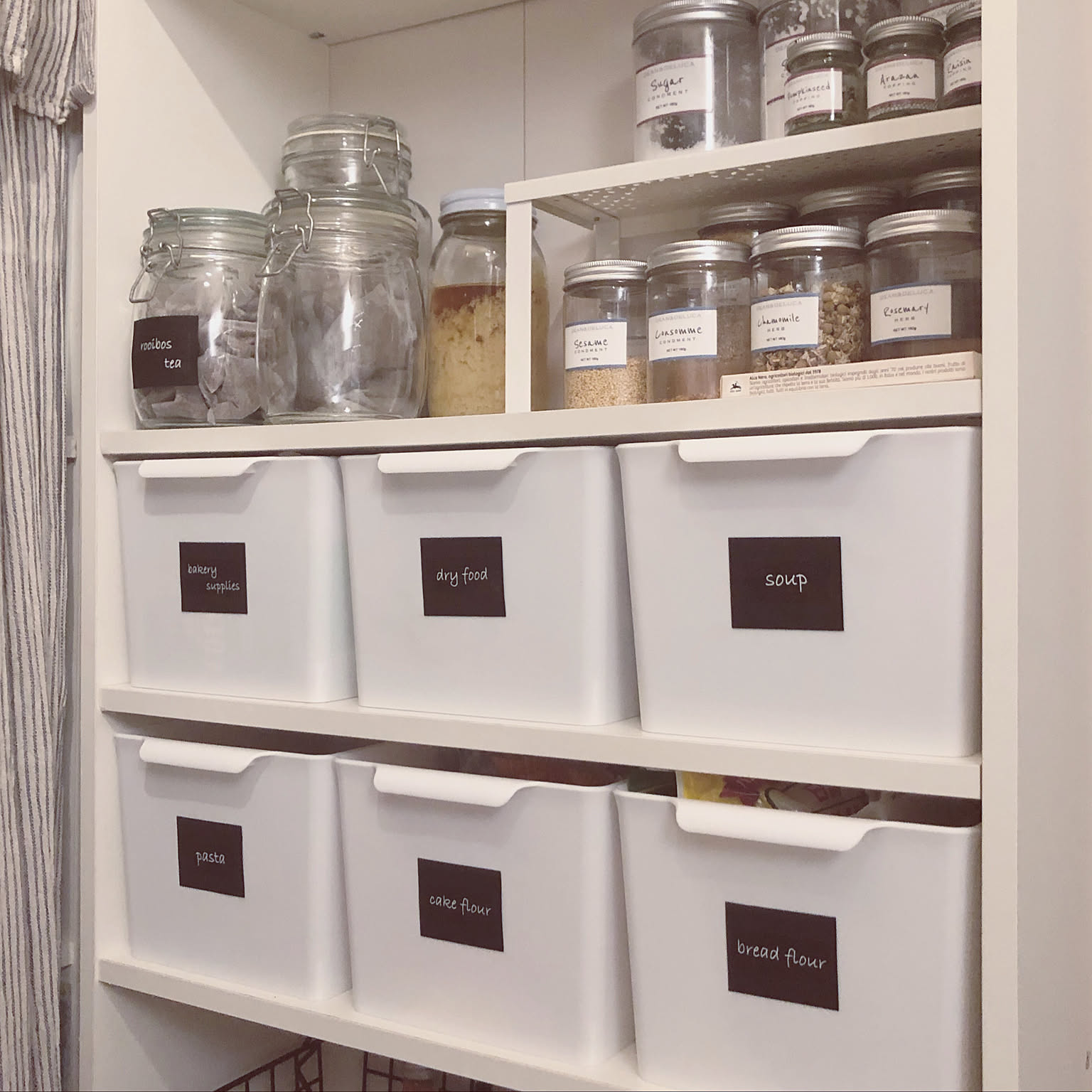 A pantry shelf with white labeled bins and clear jars for baking supplies, dry food, soup, pasta, cake flour, and bread flour.