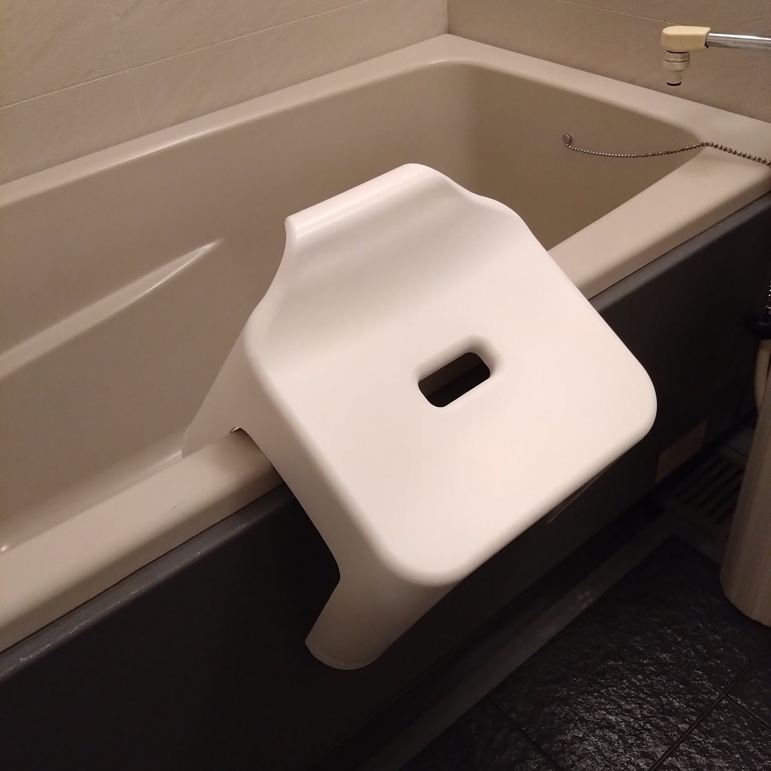 A white bath stool hooked over the edge of a bathtub so it can drip dry above the floor