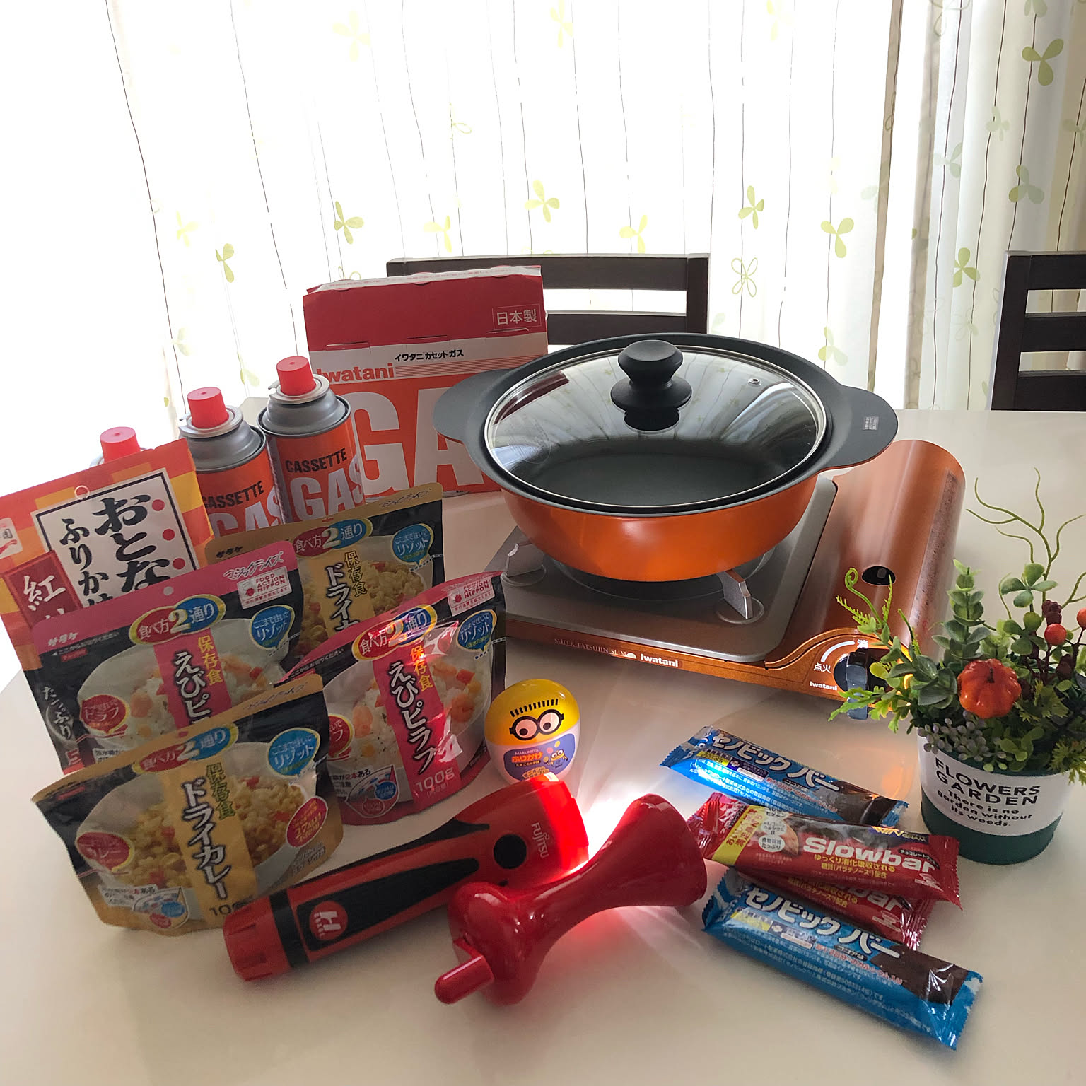 A compact Iwatani cassette stove displayed on a dining table with a pot, canned gas, and shelf-stable emergency food.