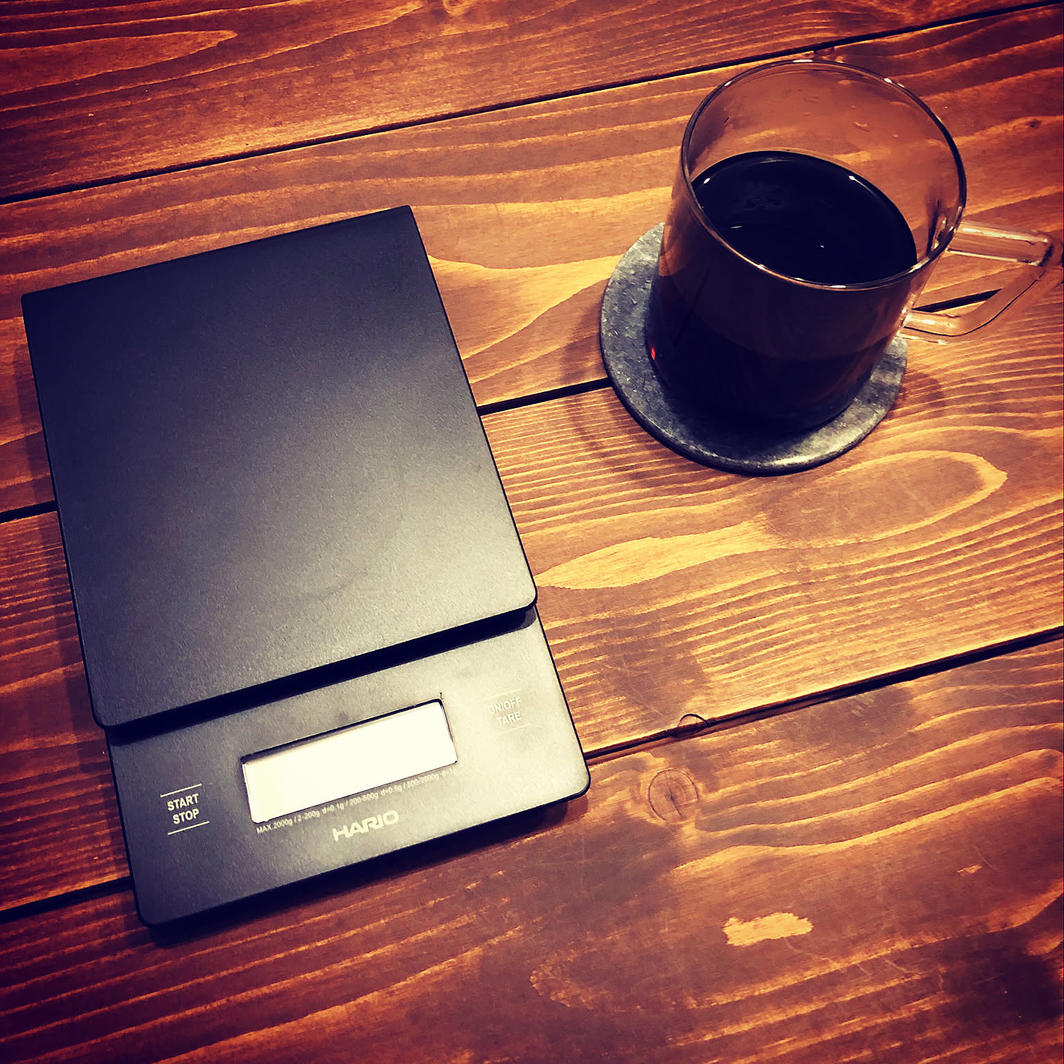 A black HARIO V60 drip scale placed beside a glass of coffee on a wooden table.