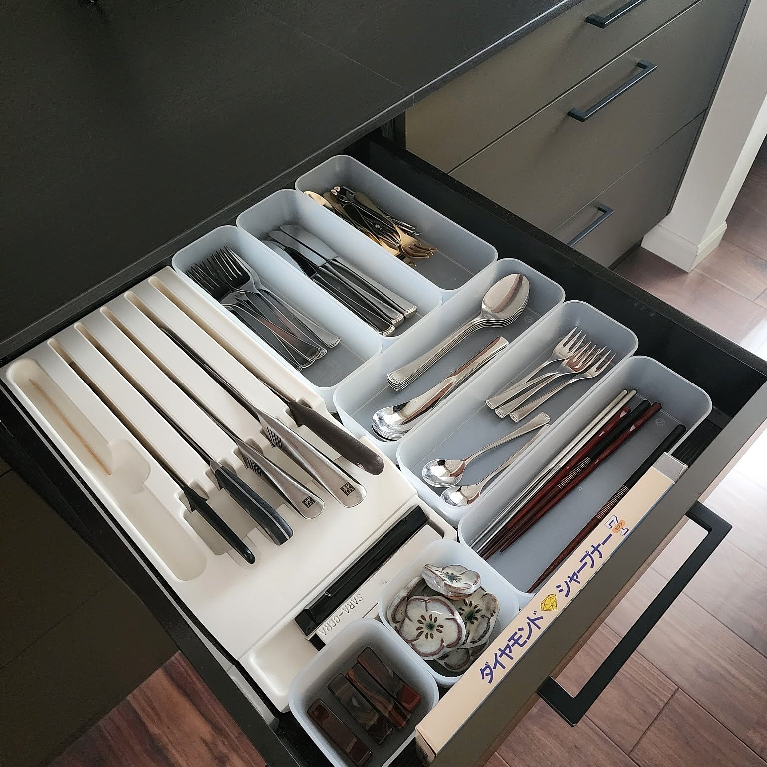 An open kitchen drawer organized with white trays for chopsticks, knives, cutlery, spoons, forks, and small daily tableware.
