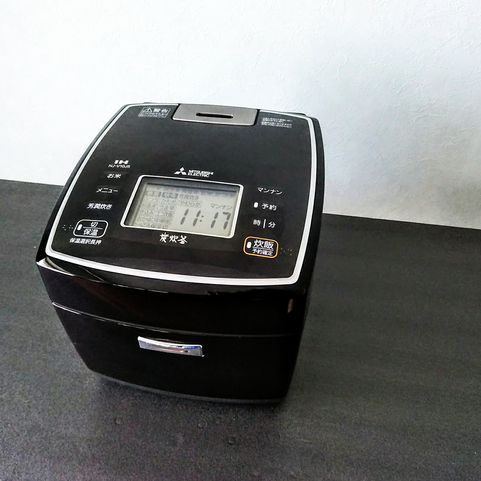 A black Mitsubishi charcoal-coated IH rice cooker with the three-diamond logo and a lit control panel, on a dark countertop