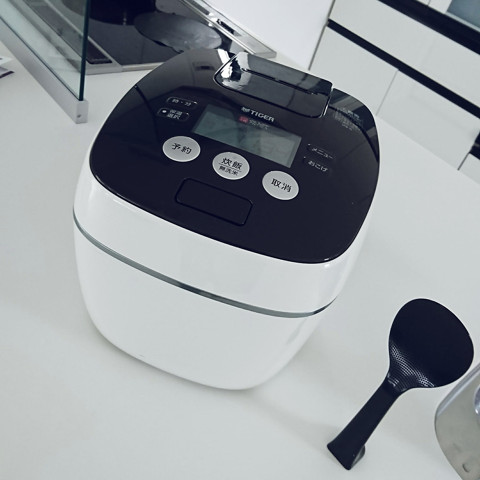 A white Tiger pressure-IH rice cooker with a glossy black lid and the TIGER logo, on a white counter with a black rice paddle