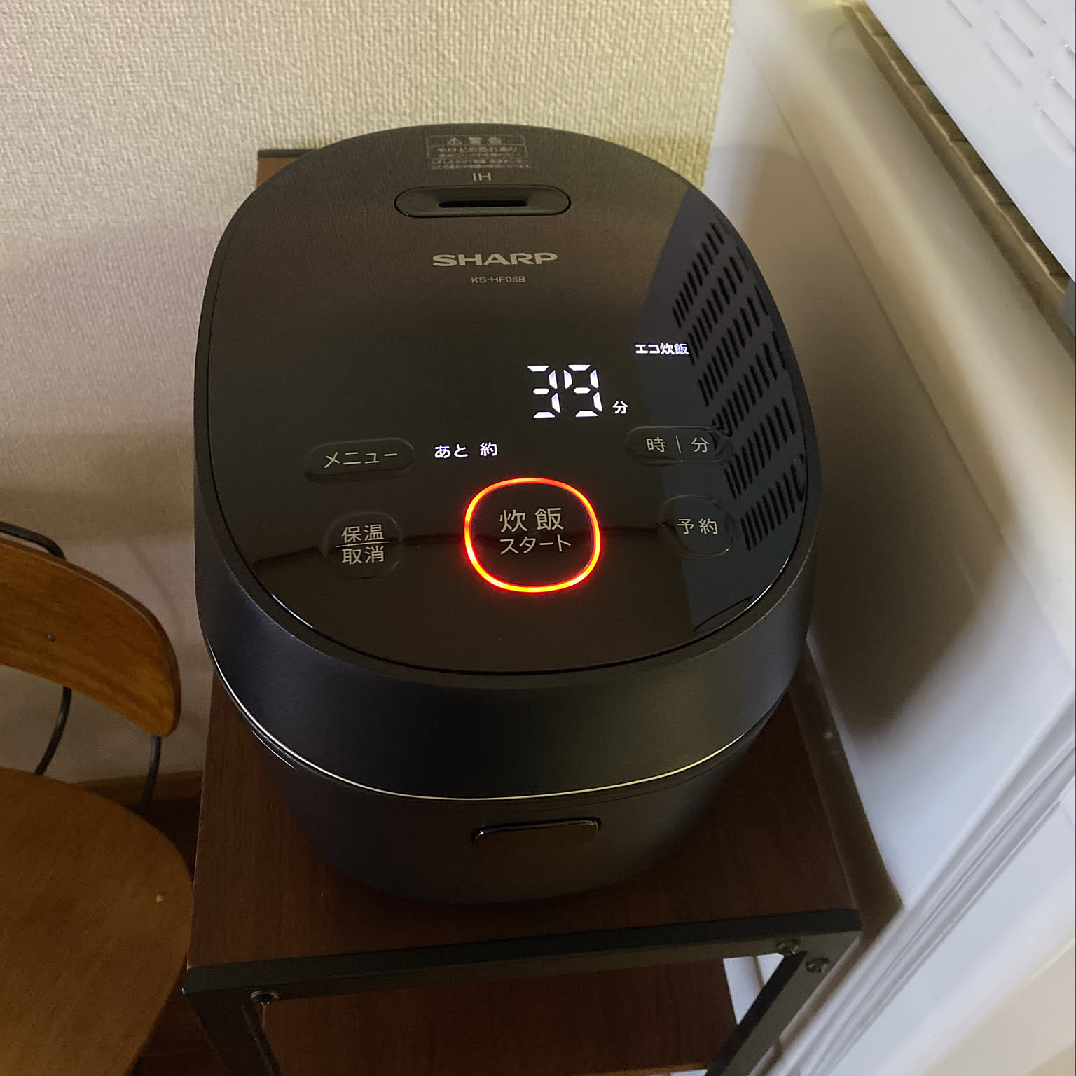 A black Sharp IH rice cooker placed on a small stand in a compact apartment.